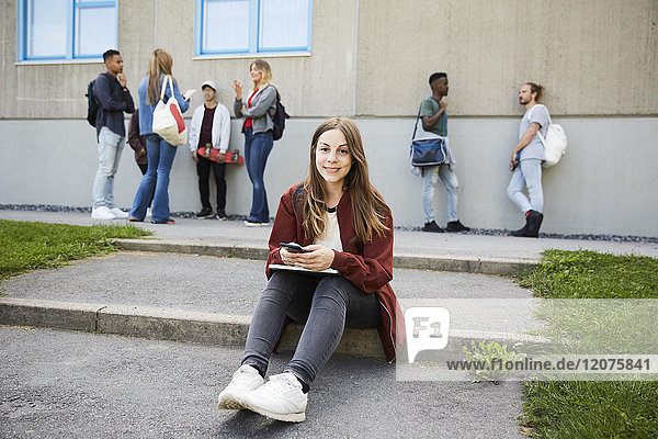 Porträt einer lächelnden Frau  die auf dem Uni-Campus mit Studenten im Hintergrund sitzt.