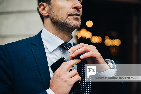 Midsection of businessman adjusting necktie while holding mobile phone