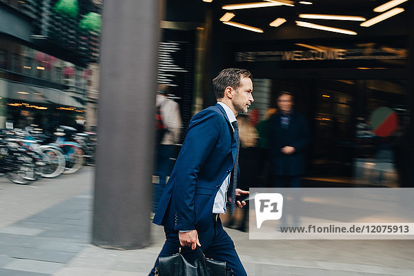 Side view of mature businessman walking with bag and smart phone on sidewalk in city
