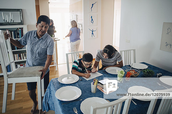 Teenage girl assisting brother in homework while father arranging chairs at dining table