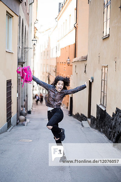 Volle Länge der aufgeregten Teenagerin beim Springen mit Rucksack auf der Straße inmitten von Gebäuden.
