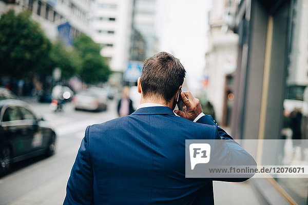 Rear view of mature businessman talking on mobile phone while walking on sidewalk in city