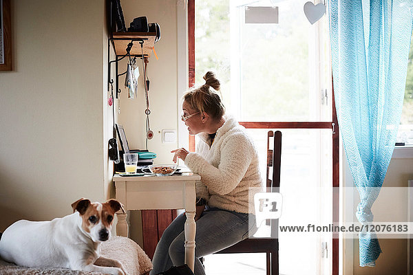 Behinderte junge Frau mit Laptop am Schreibtisch mit Hund im Vordergrund zu Hause