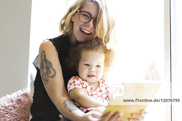 Mother sitting with daughter (12-17 months) on windowsill and reading book