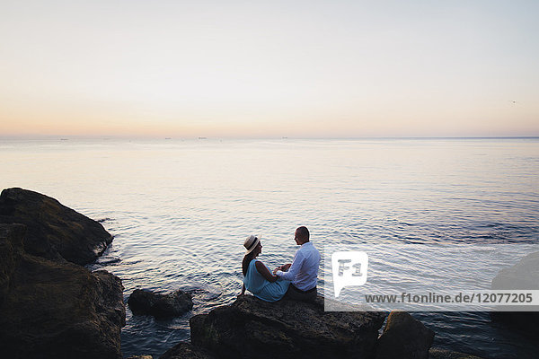 Kaukasisches Paar sitzt bei Sonnenuntergang auf einem Felsen am Meer