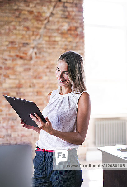 Smiling businesswoman holding clipboard at brick wall in office