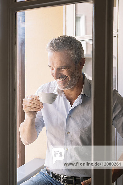 Lachender  reifer Mann trinkt eine Tasse Kaffee am Fenster.