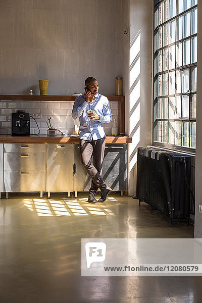 Young entrepreneur standing in company kitchen  drinking coffee  using smartphone