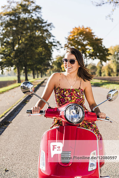 Glückliche junge Frau auf dem Motorroller auf der Landstraße