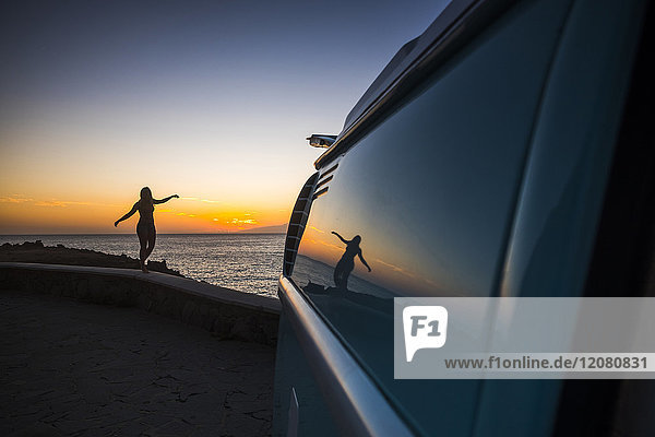Silhouette einer Frau die bei Sonnenuntergang an der Wand vor dem Meer balanciert.