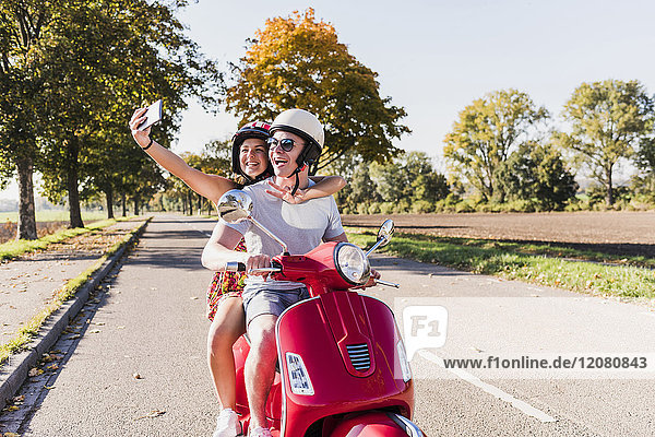 Glückliches junges Paar mit einem Selfie auf dem Motorroller auf der Landstraße