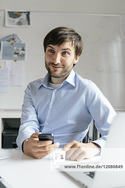 Smiling businessman with cell phone and laptop at desk in office