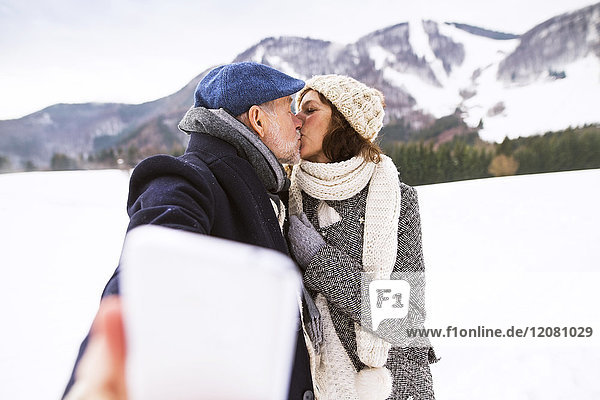 Senior couple kissing in winter landscape while taking selfie with cell phone