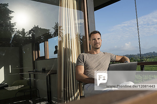 Mann  der auf dem Balkon eines modernen Hauses sitzt und einen Laptop hält.