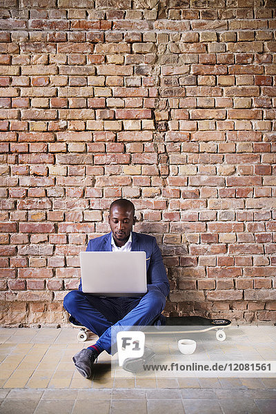 Businessman sitting on longboard at brick wall using laptop