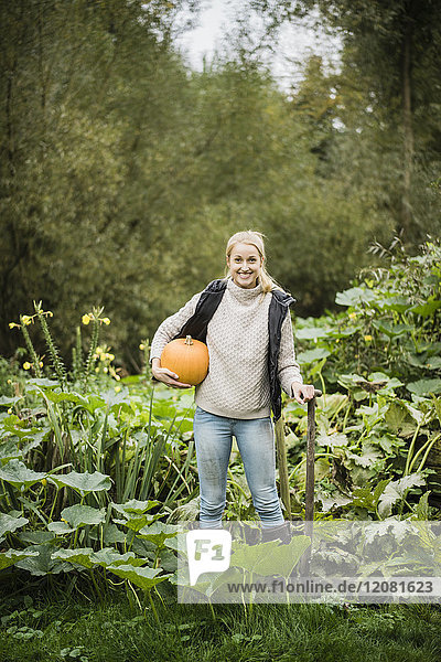 Porträt der lächelnden jungen Frau mit Spaten und Kürbis im Garten