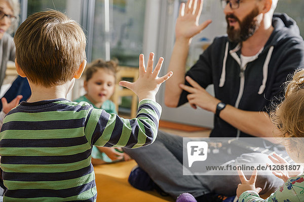 Child raising hand while singing with others and teacher in kindergarten
