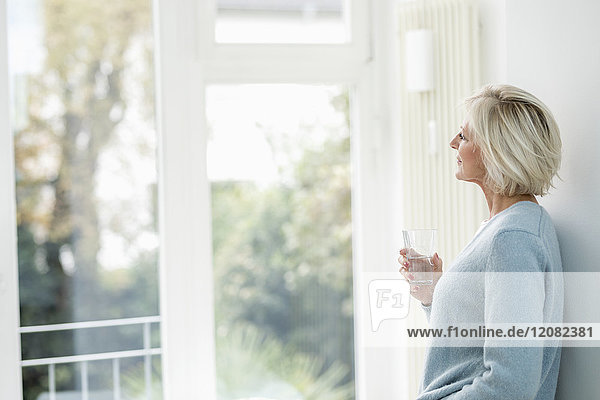 Lächelnde Seniorin mit einem Glas Wasser aus dem Fenster schauend