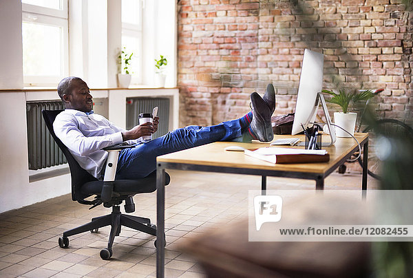 Geschäftsmann im Büro sitzend mit Füßen auf dem Schreibtisch beim Handy-Checken