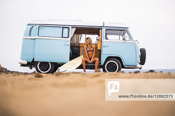 Junge Frau mit Surfbrett im Van am Strand sitzend