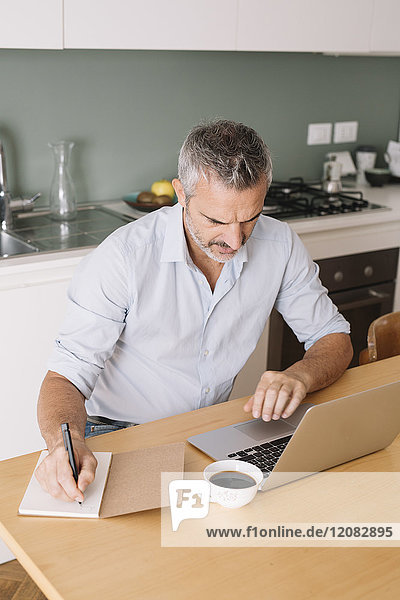 Mann schreibt in Notizbuch und benutzt Laptop im Home-Office