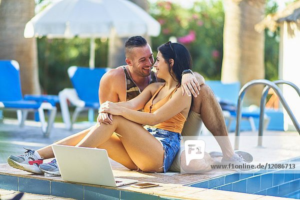Young couple laughing together while sitting with mobile laptop next to pool in holiday resort. Greek ethnicity. In Hersonissos  Crete  Greece