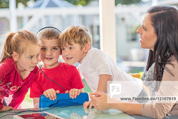 Mittlere erwachsene Frau sitzt mit drei kleinen Kindern  ein Junge hält ein digitales Tablet und trägt Kopfhörer