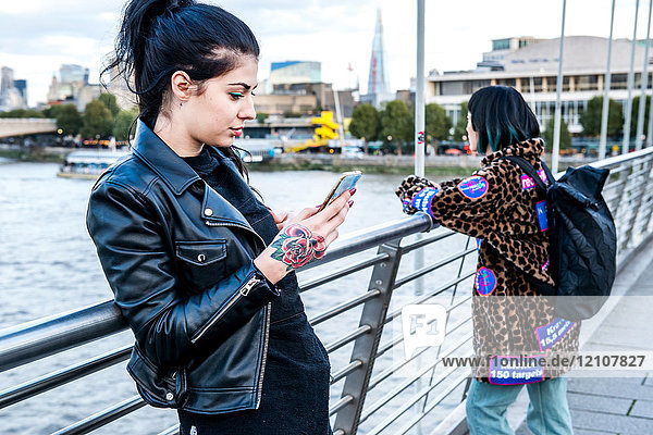 Junge Frau betrachtet Smartphone mit Freundin  die von der Millennium-Fußgängerbrücke hinaussieht  London  Großbritannien