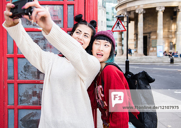 Two young stylish women taking selfie by red phone box  London  UK
