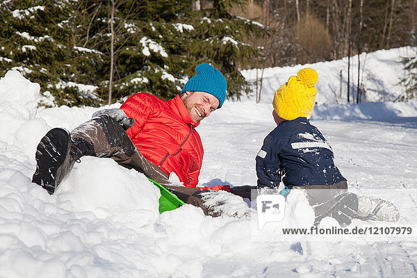 Man and son laughing after falling from toboggan in snow covered landscape