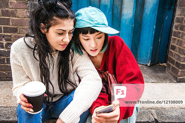 Zwei junge Frauen sitzen auf dem Bürgersteig und schauen auf ein Smartphone