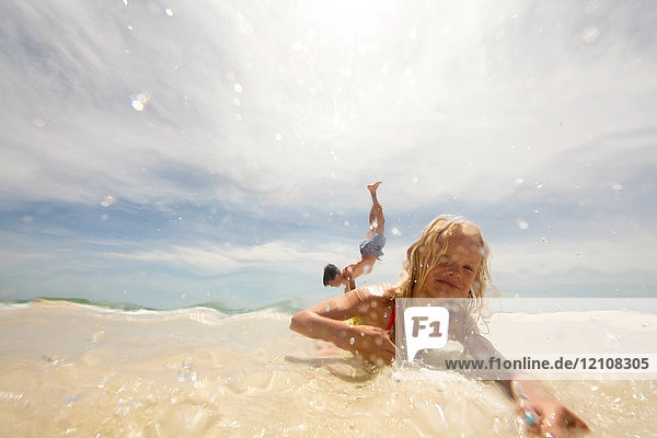 Mädchen liegt am Strand im flachen Wasser  Bruder macht Handstand im Hintergrund