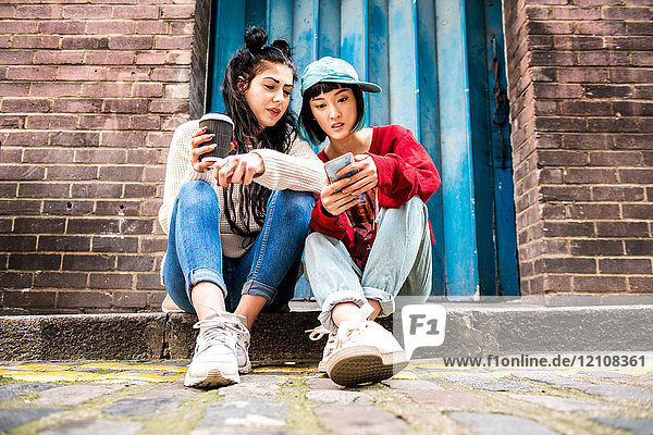 Zwei junge Frauen sitzen auf dem Bürgersteig und schauen auf ein Smartphone