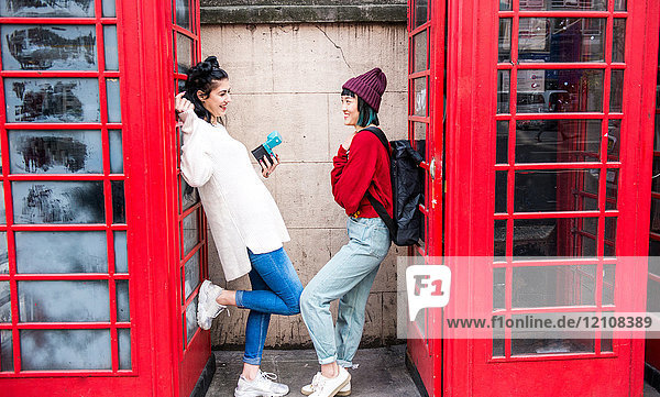 Two young stylish women leaning against red phone boxes  London  UK