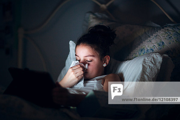 Mädchen im Bett schnäuzt sich die Nase  beleuchtet vom Licht des digitalen Tabletts