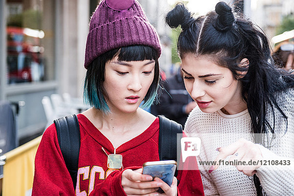 Zwei junge  stilvolle Frauen schauen sich auf der Straße in der Stadt ein Smartphone an
