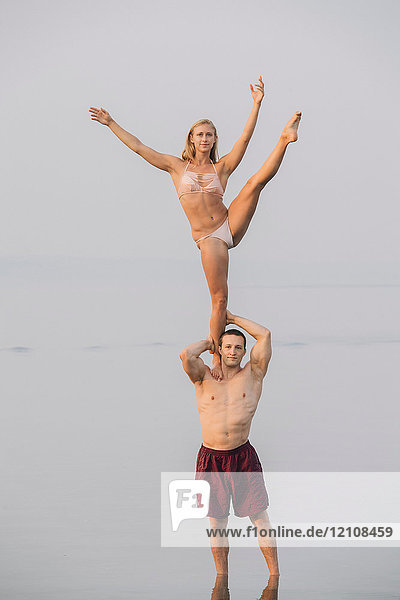 Junges Paar am Strand  Frau balanciert auf einem Bein  auf der Schulter eines Mannes