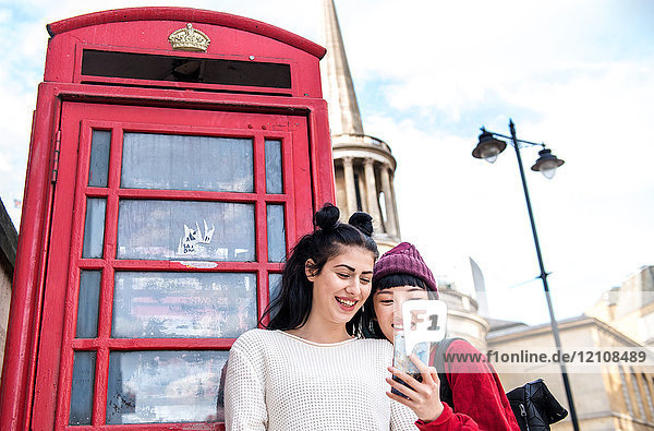 Zwei junge  stilvolle Frauen betrachten ein Smartphone an einer roten Telefonzelle  London  UK