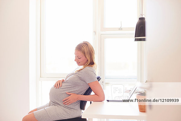 Schwangere junge Frau am Schreibtisch mit Blick auf den Bauch