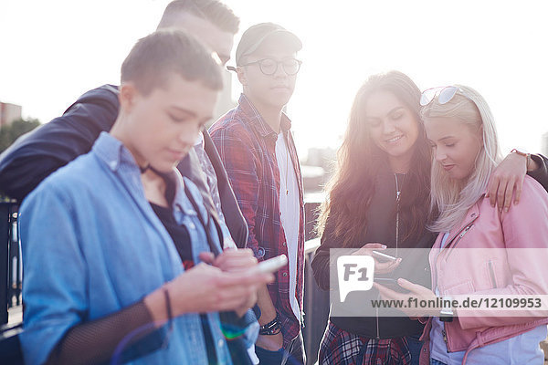 Fünf junge erwachsene Freunde schauen sich in der sonnenbeschienenen Stadt Smartphones an