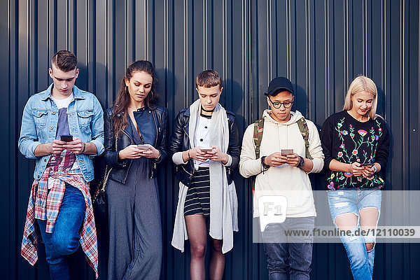 Row of five young adult friends leaning against black wall looking at smartphones