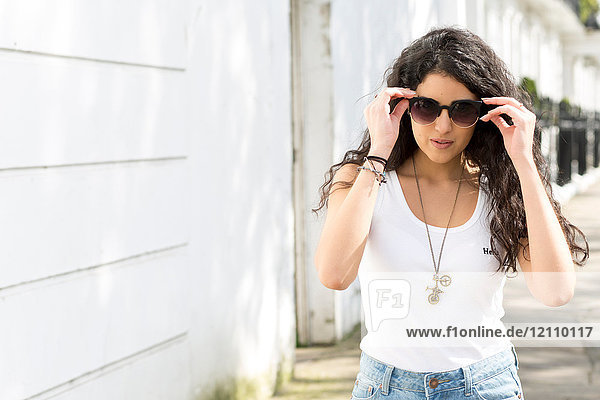 Junge Frau mit langen gewellten Haaren schlendert auf der Straße und setzt eine Sonnenbrille auf