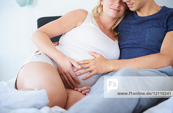 Schnappschuss eines Mannes und einer schwangeren Freundin  die mit den Händen auf dem Bauch im Bett sitzen