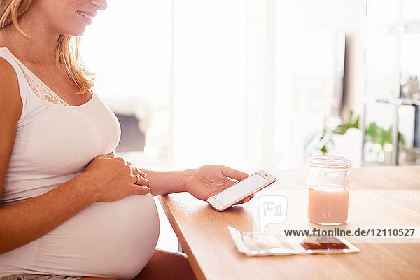 Ausschnittaufnahme einer schwangeren jungen Frau am Schreibtisch beim Betrachten von Smartphone- und Ultraschallbildern