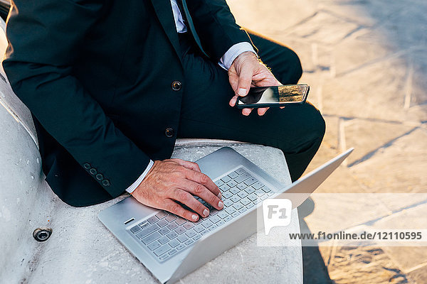 Reifer Geschäftsmann sitzt im Freien  benutzt Laptop  hält Smartphone  niedriger Abschnitt