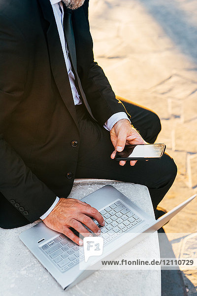 Reifer Geschäftsmann sitzt im Freien  benutzt Laptop  hält Smartphone  Mittelteil