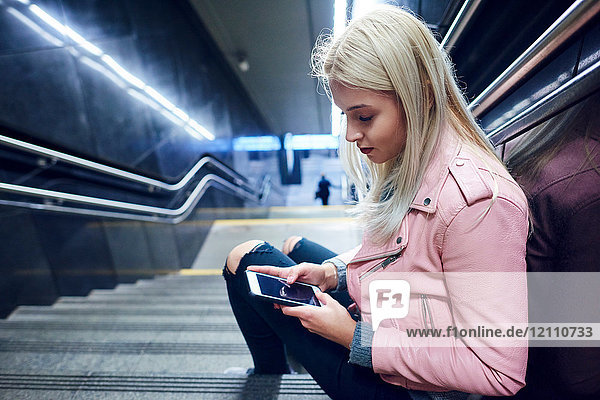 Junge Frau sitzt auf der Treppe der U-Bahn-Station und schaut auf ein Smartphone