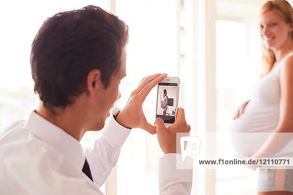 Over shoulder view of man taking smartphone photograph of pregnant girlfriend