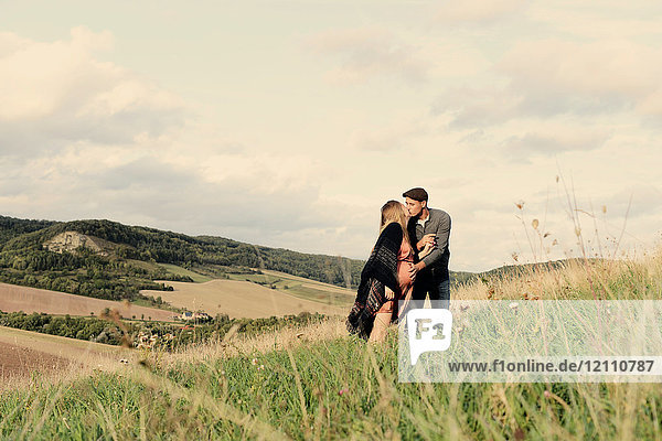 Romantic pregnant mid adult couple kissing on rural hillside