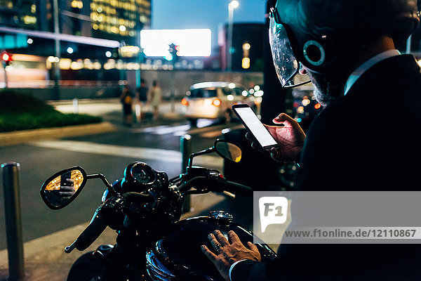 Älterer Geschäftsmann nachts im Freien  auf dem Motorrad sitzend  Motorradhelm tragend  Smartphone benutzen  Rückansicht